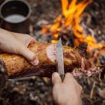 Man carving grilled rabbit meat in forest camp