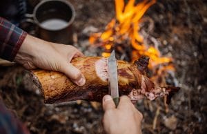 man carving grilled rabbit meat in forest camp