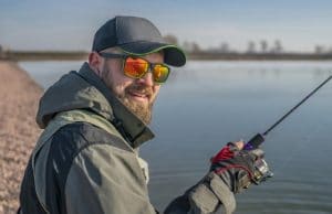 Portrait of fisherman. Bearded man in cap and sunglasses holds fishing rod at lake