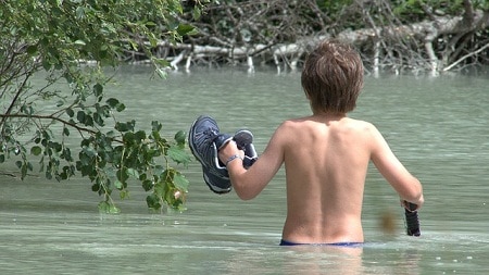 kid holding his shoes while crossing a river