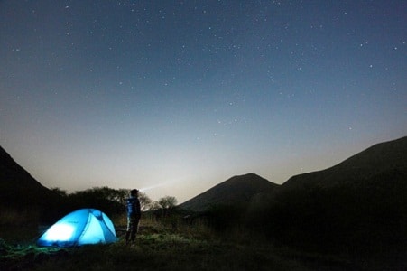 man standing staring at a starry sky by a lit tent