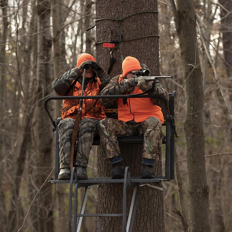 Two people sitting in a treestand hunting, one with binoculars and one with aiming rifle