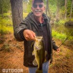 Man holding a bass in one hand and fishing rod in the other in the fall