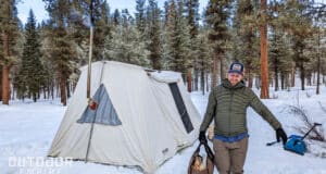 Man carrying firewood in a tote in front of a hot tent in the snow while winter camping