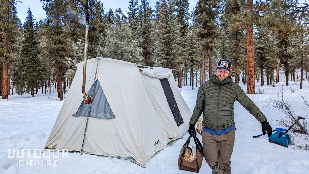 Man carrying firewood in a tote in front of a hot tent in the snow while winter camping