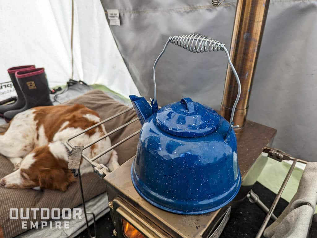 Blue enamel teak kettle from GSI Outdoors sitting on a wood stove in a tent with dog sleeping next to it