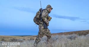 Hunter walking through dry grass with rifle on shoulder and wearing Sitka camouflage clothing