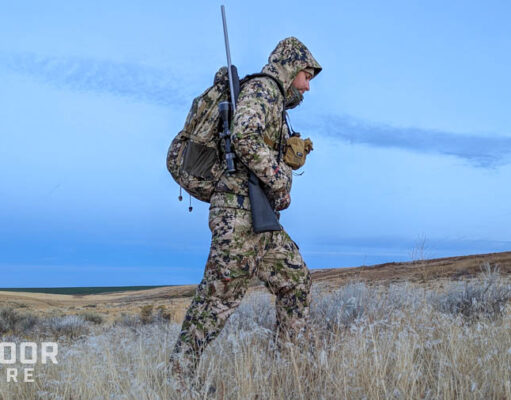 Hunter walking through dry grass with rifle on shoulder and wearing Sitka camouflage clothing