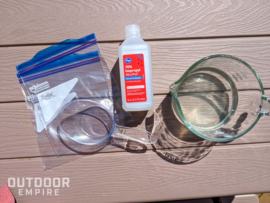 Materials for making a gel ice pack sitting on a table including ziploc bags, measuring cup, rubbing alcohol and pitcher of water