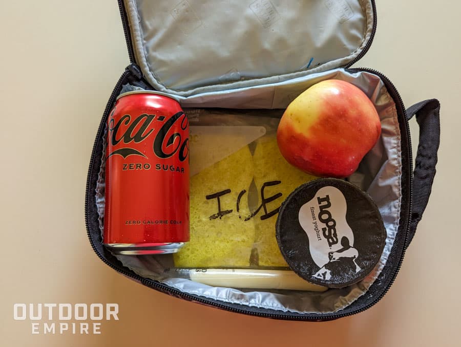 An open lunch box with a DIY ice pack inside as well as apple, yogurt, cheese, and a Coke