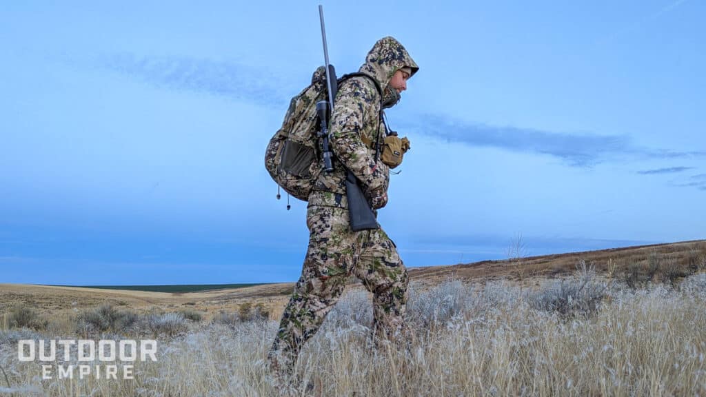 Hunter in camo walking through field in desert with rifle on shoulder